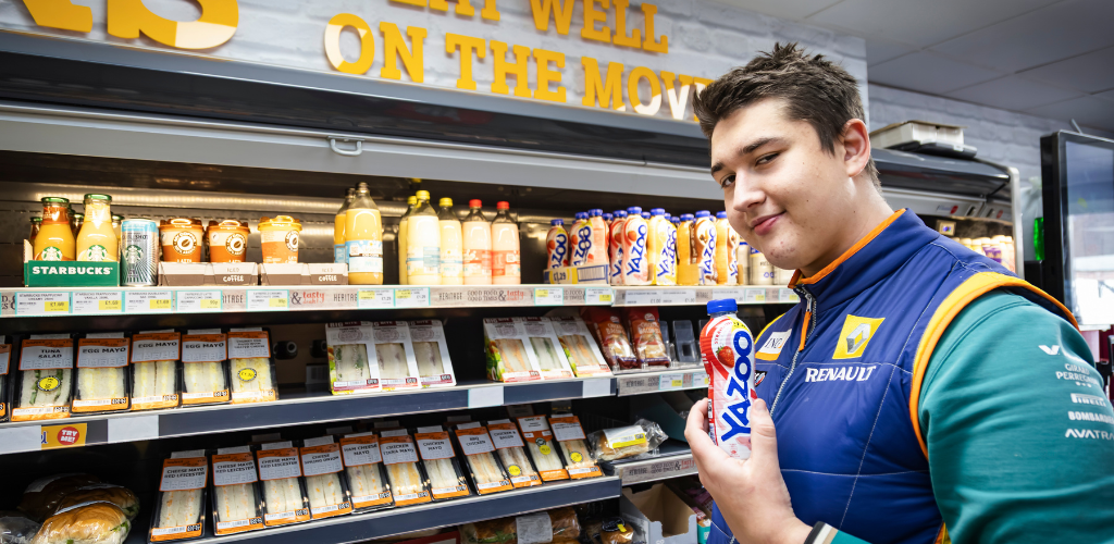 a student in a grocery store, smiling at the camera and holding up a Yazoo bottle